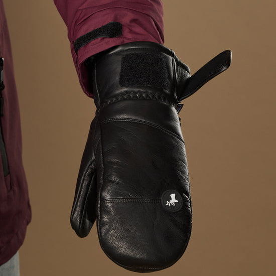 Black leather mitten close-up showing a wrist strap, stitched cuff, and logo patch.